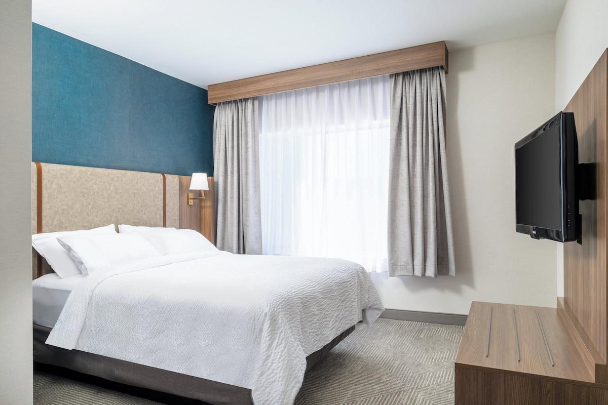 Een hotelkamer met een bed, een aan de muur bevestigde televisie, een raam met gordijnen en een houten hoofdeinde.