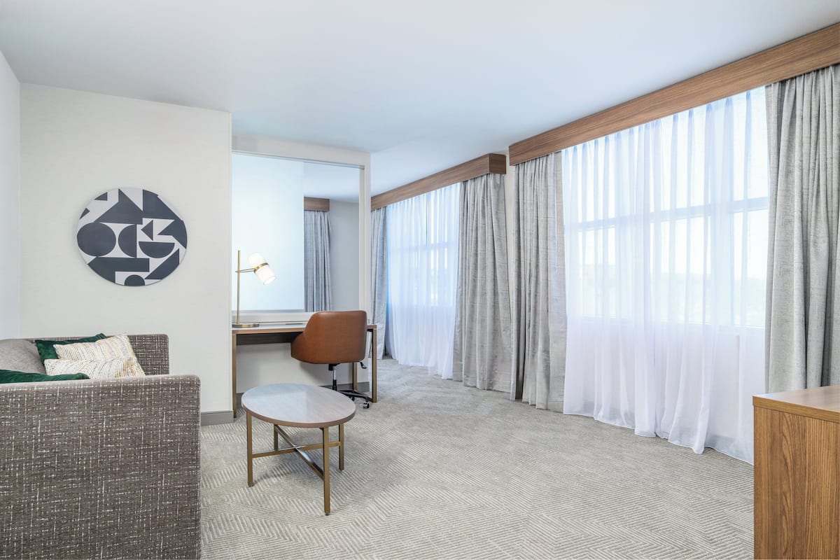 Une chambre d’hôtel avec un canapé, une petite table ronde, une chaise, un bureau et une grande fenêtre avec des rideaux légers.