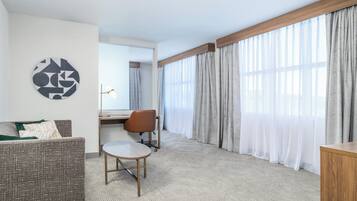 Habitación de hotel con sofá, una mesita redonda pequeña, una silla, un escritorio y un ventanal con cortinas translúcidas.