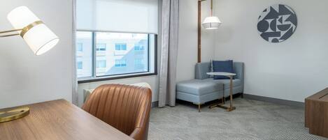 Habitación de hotel con un escritorio de madera, una silla de cuero, un sillón pequeño, una mesa redonda y un gran ventanal con cortinas.