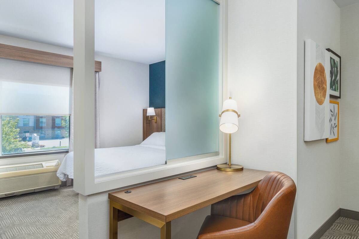 Une chambre d’hôtel comprenant un lit, un bureau, une chaise, une lampe et une fenêtre donnant sur un immeuble.