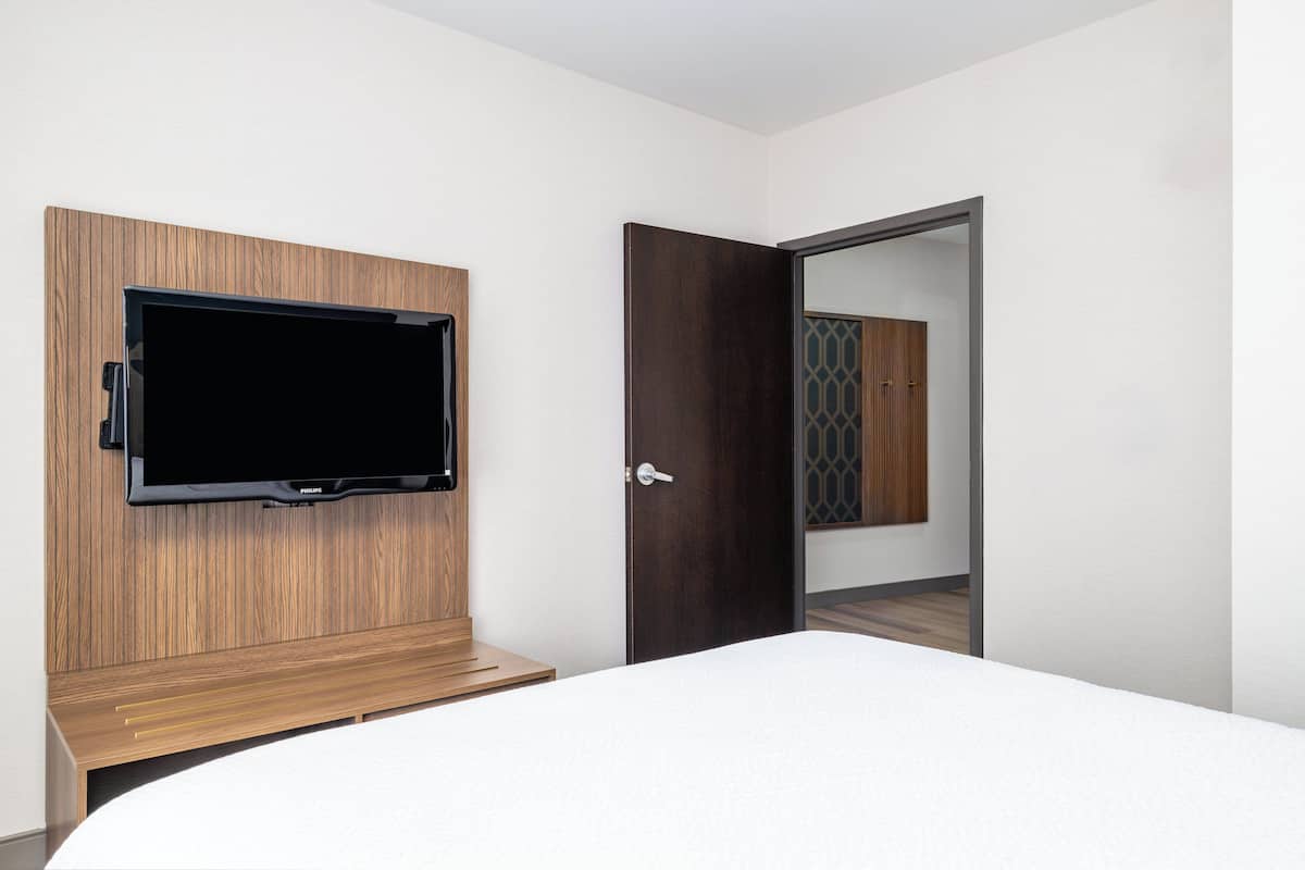 Une chambre d’hôtel moderne dotée d’un téléviseur à écran plat fixé sur un panneau en bois, d’un lit avec une literie blanche et d’une porte donnant sur une autre pièce dont le mur est décoré d’un motif.