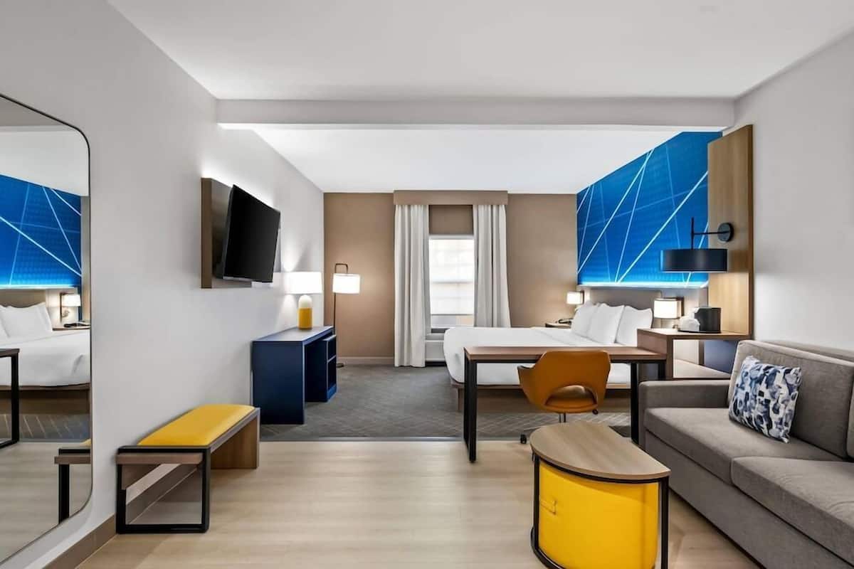 A hotel room with a bed, a desk, a chair, a sofa, a TV, and a mirror.