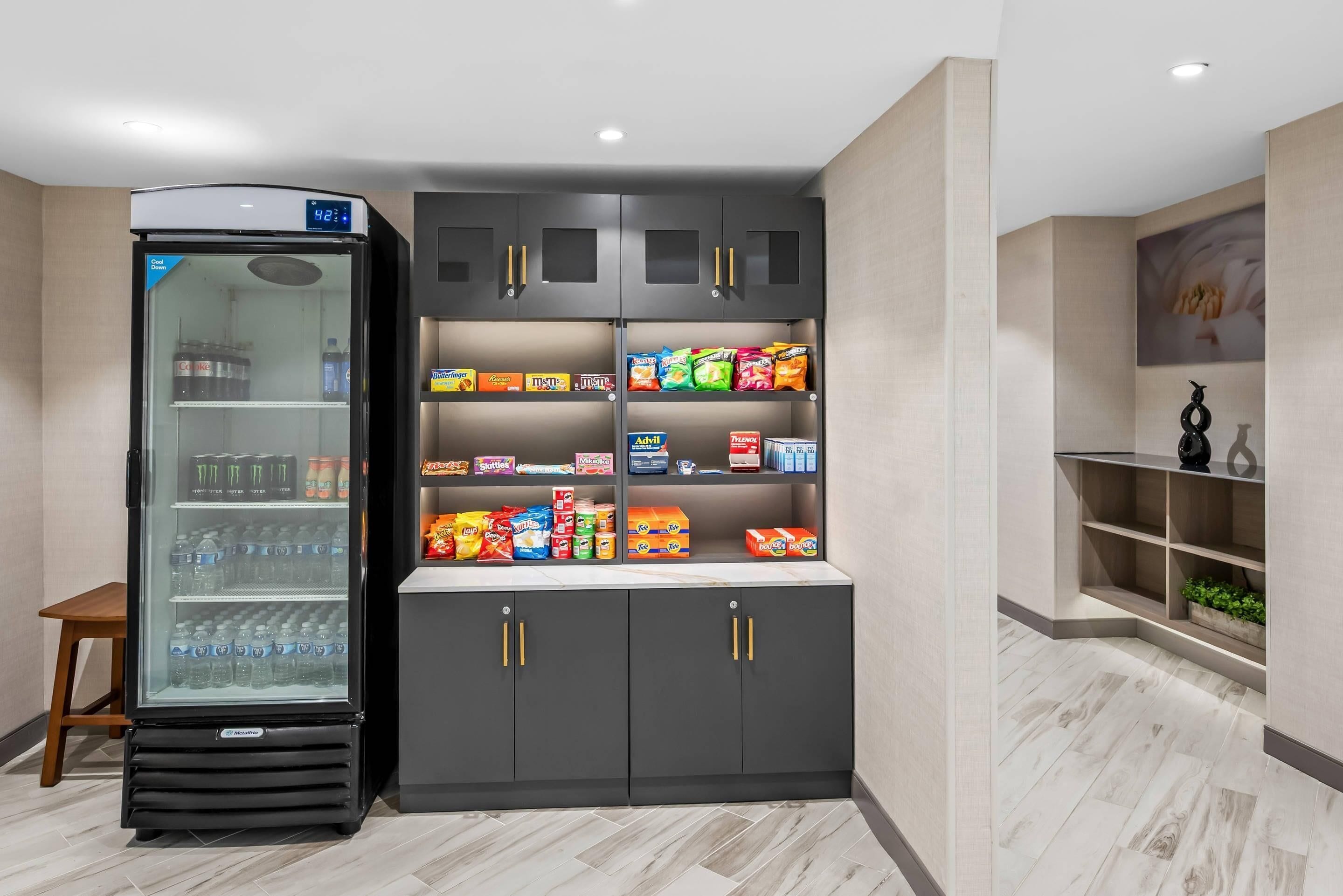 A modern kitchenette with a refrigerator, shelves stocked with snacks, and a small seating area.