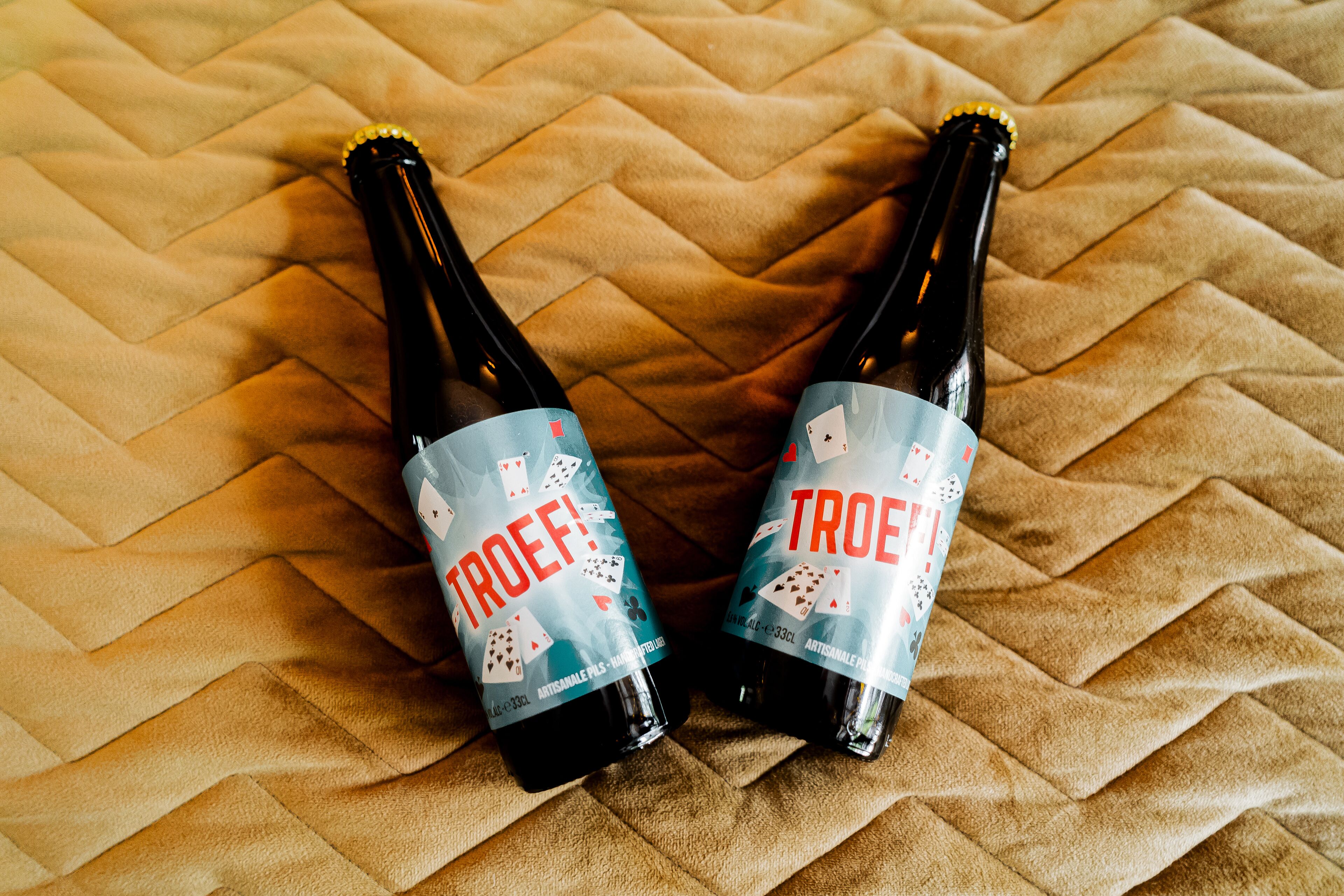 Two bottles of Troef beer with a card game theme on a textured fabric surface.