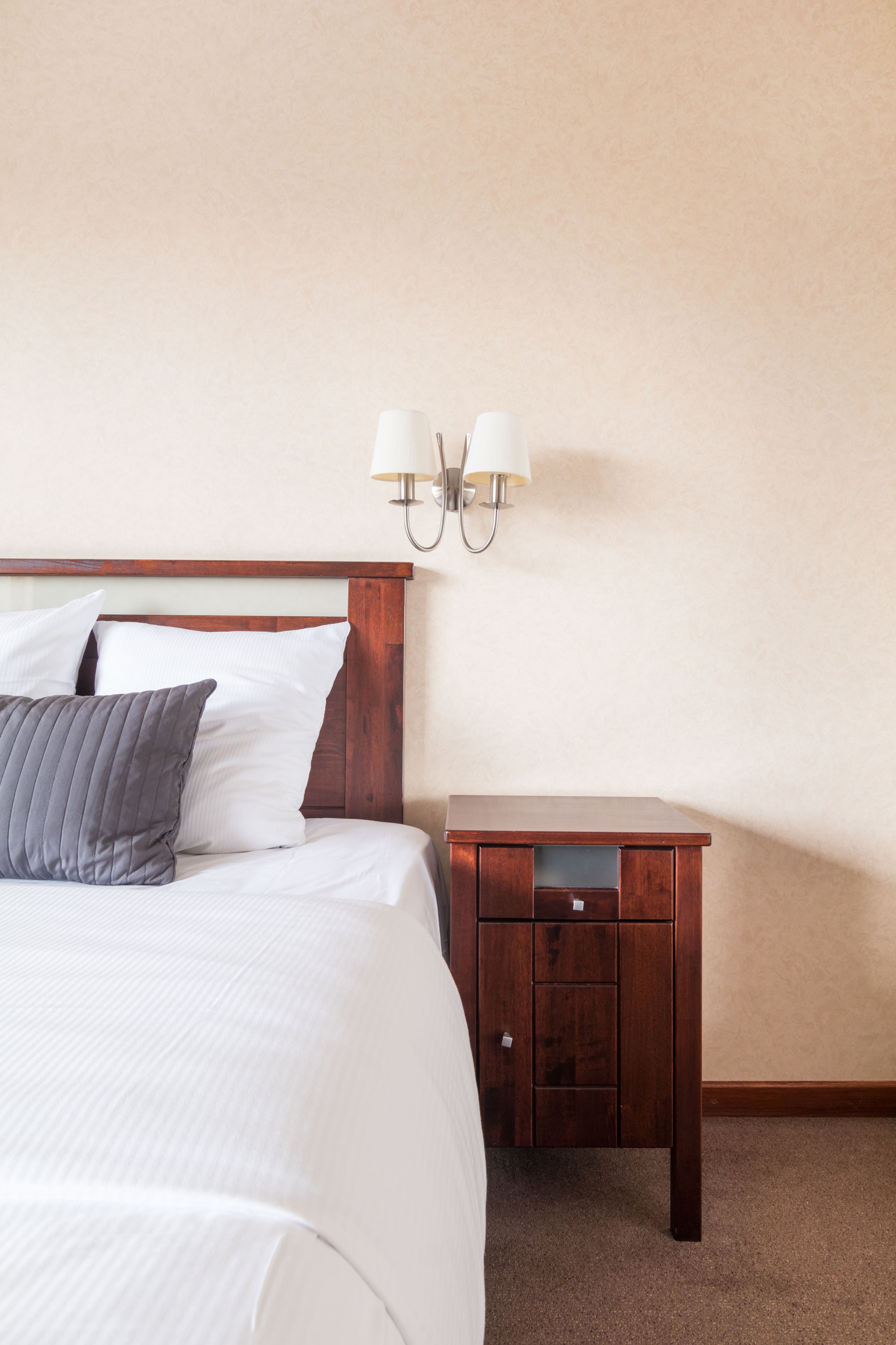 Una cama bien hecha con almohadas blancas y grises, una mesita de noche de madera con cajones y una lámpara fijada a la pared.