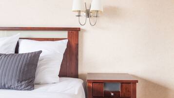 Una cama bien hecha con almohadas blancas y grises, una mesita de noche de madera con cajones y una lámpara fijada a la pared.