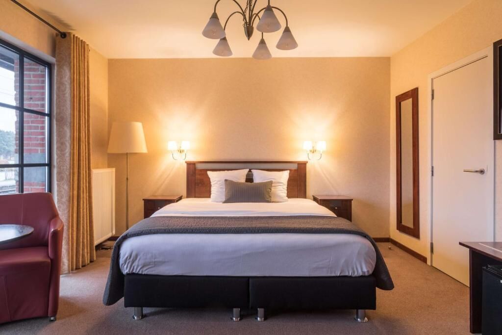 A hotel room with a large bed, two bedside lamps, a red armchair, a small table, and a window with curtains.