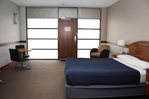A hotel room with a large bed, a chair, a desk, and a window with blinds.