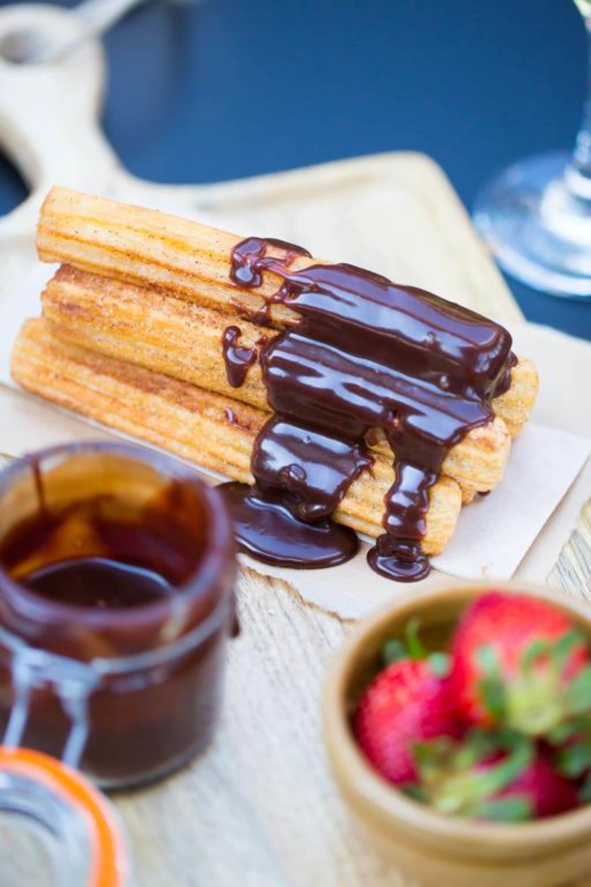 A stack of churros with chocolate sauce on a wooden board, accompanied by a jar of chocolate sauce and a bowl of strawberries.
