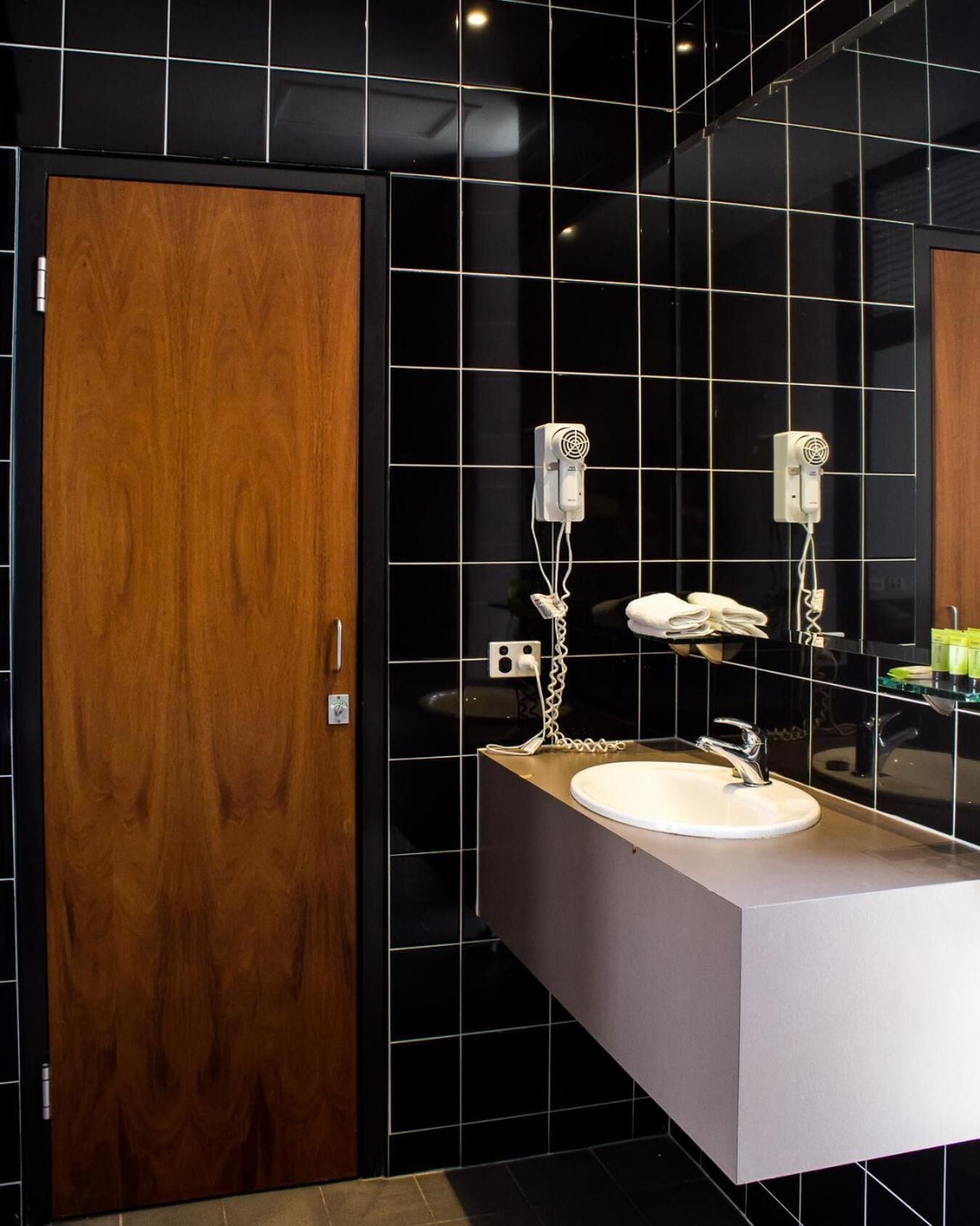Une salle de bain moderne avec une porte en bois, des murs recouverts de carreaux noirs, un lavabo blanc et deux sèche-cheveux fixés au mur.