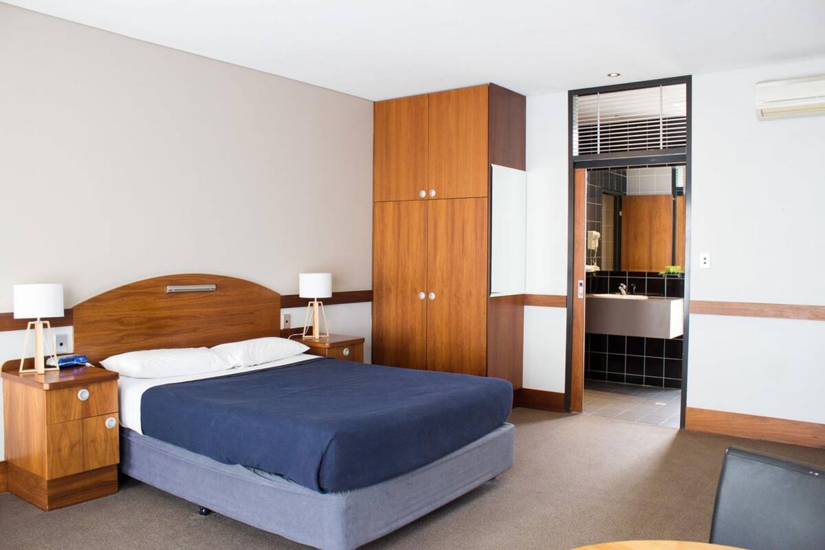 A hotel room with a bed, wooden wardrobe, bedside tables, and a bathroom with a sink and mirror.