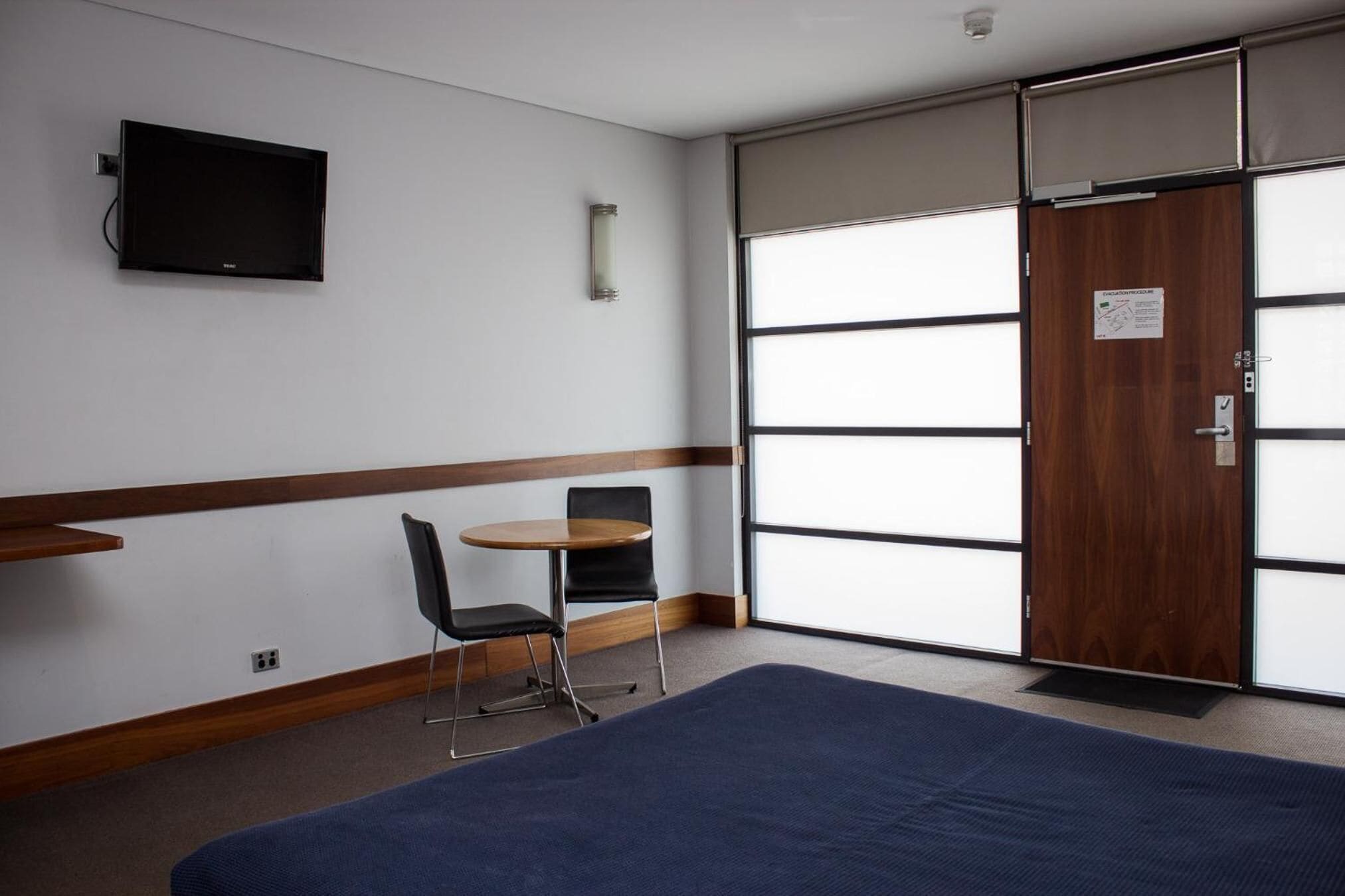 Habitación de hotel con cama, escritorio, dos sillas, televisor montado en la pared y un ventanal con puerta corrediza.