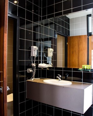 A modern bathroom with a black tiled wall, a large mirror, a white sink, and two hair dryers mounted on the wall. - Highlander Hotel (Gilles Plains)