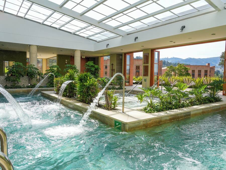 An indoor pool with multiple water features, surrounded by greenery and with a view of buildings and mountains in the background.