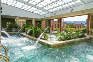 Una piscina interior con múltiples elementos acuáticos, rodeada de follaje y con vistas a edificios y montañas al fondo.