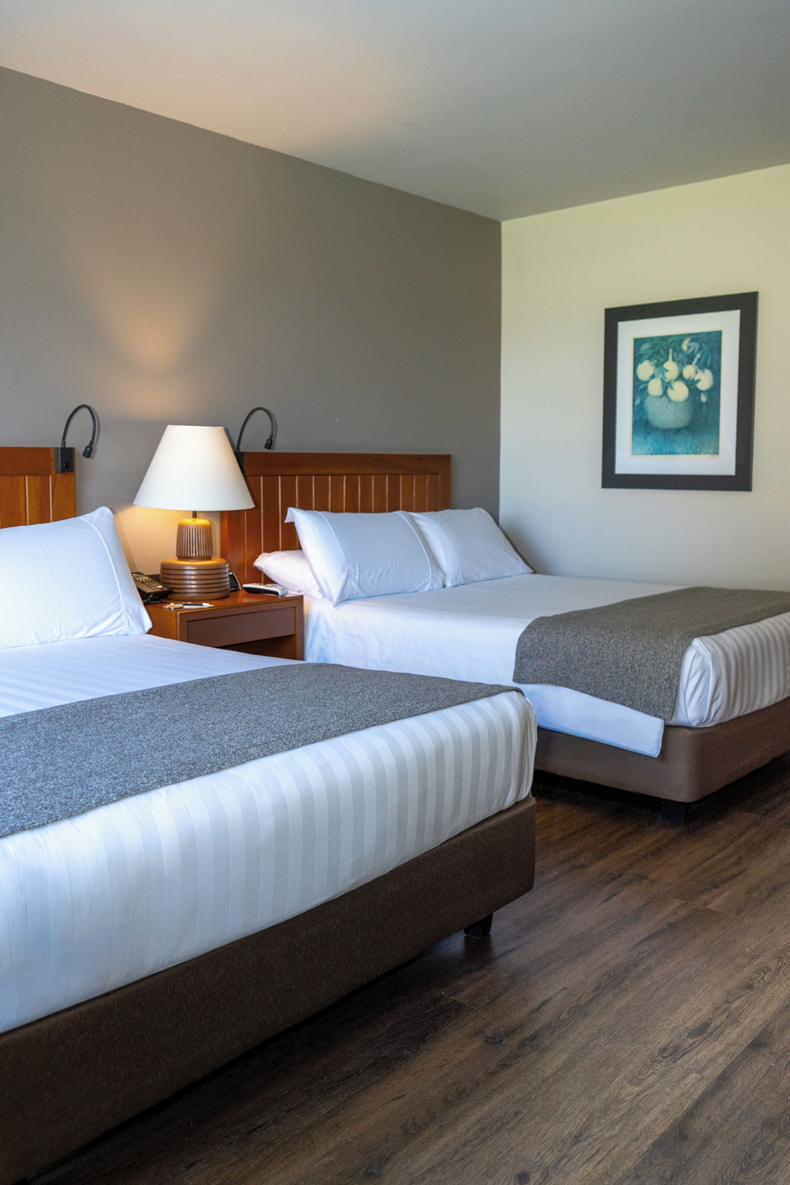 Quarto de hotel com duas camas, uma mesinha de cabeceira com abajur, um quadro pendurado na parede e piso de madeira.