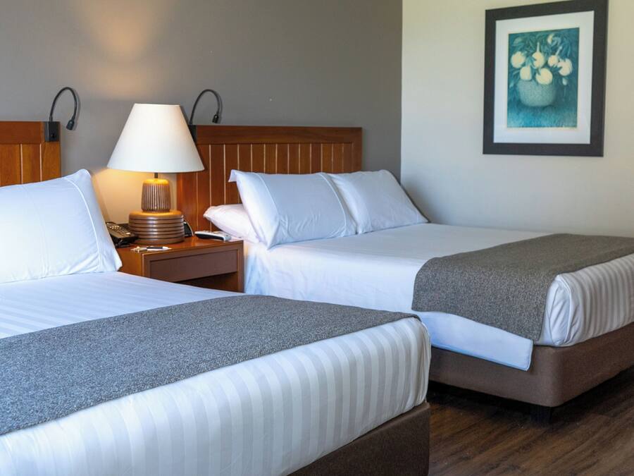 Quarto de hotel com duas camas, uma mesinha de cabeceira com abajur, um quadro pendurado na parede e piso de madeira.