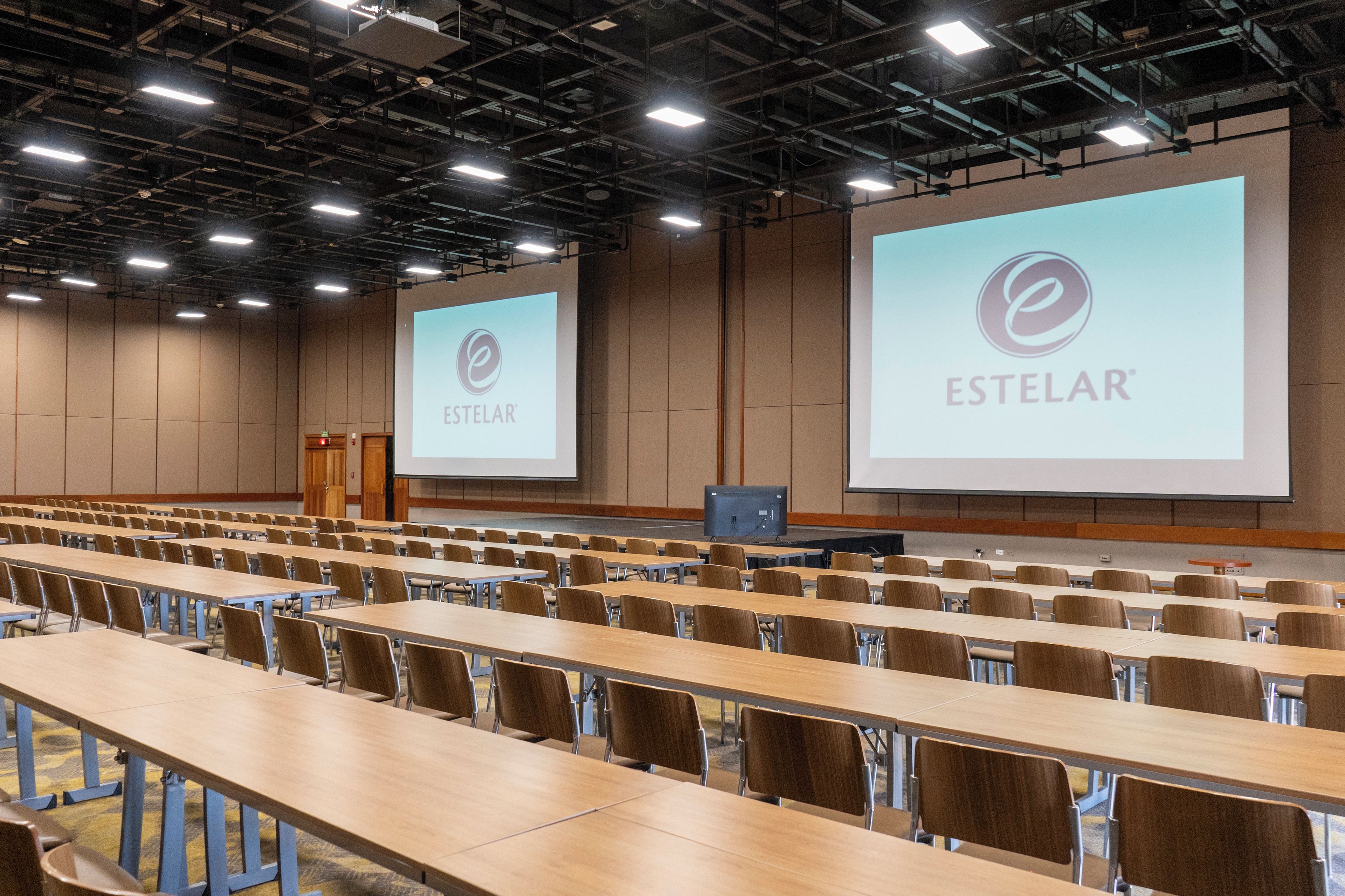 Un salón de conferencias con filas de mesas y sillas, grandes pantallas de proyección y un escenario con un atril.