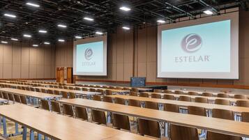 Un salón de conferencias con filas de mesas y sillas, grandes pantallas de proyección y un escenario con un atril.
