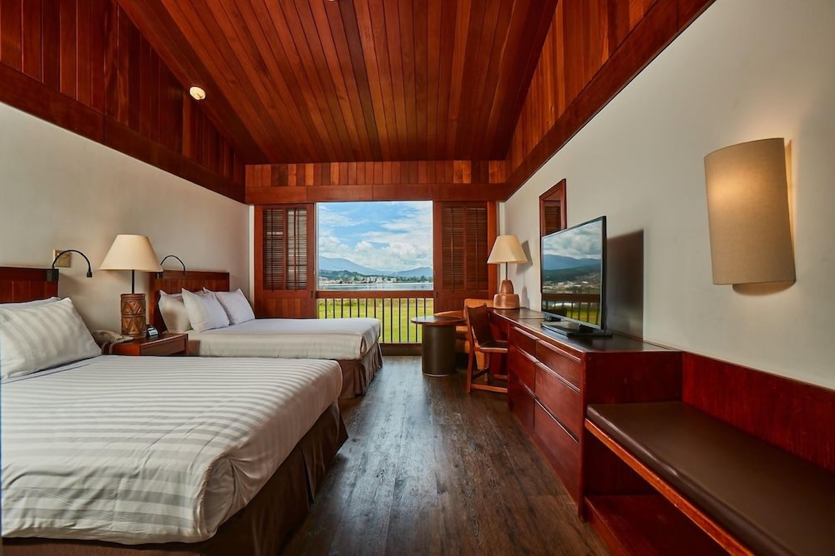 A hotel room with two beds, a television, a desk, and a view of the outdoors.
