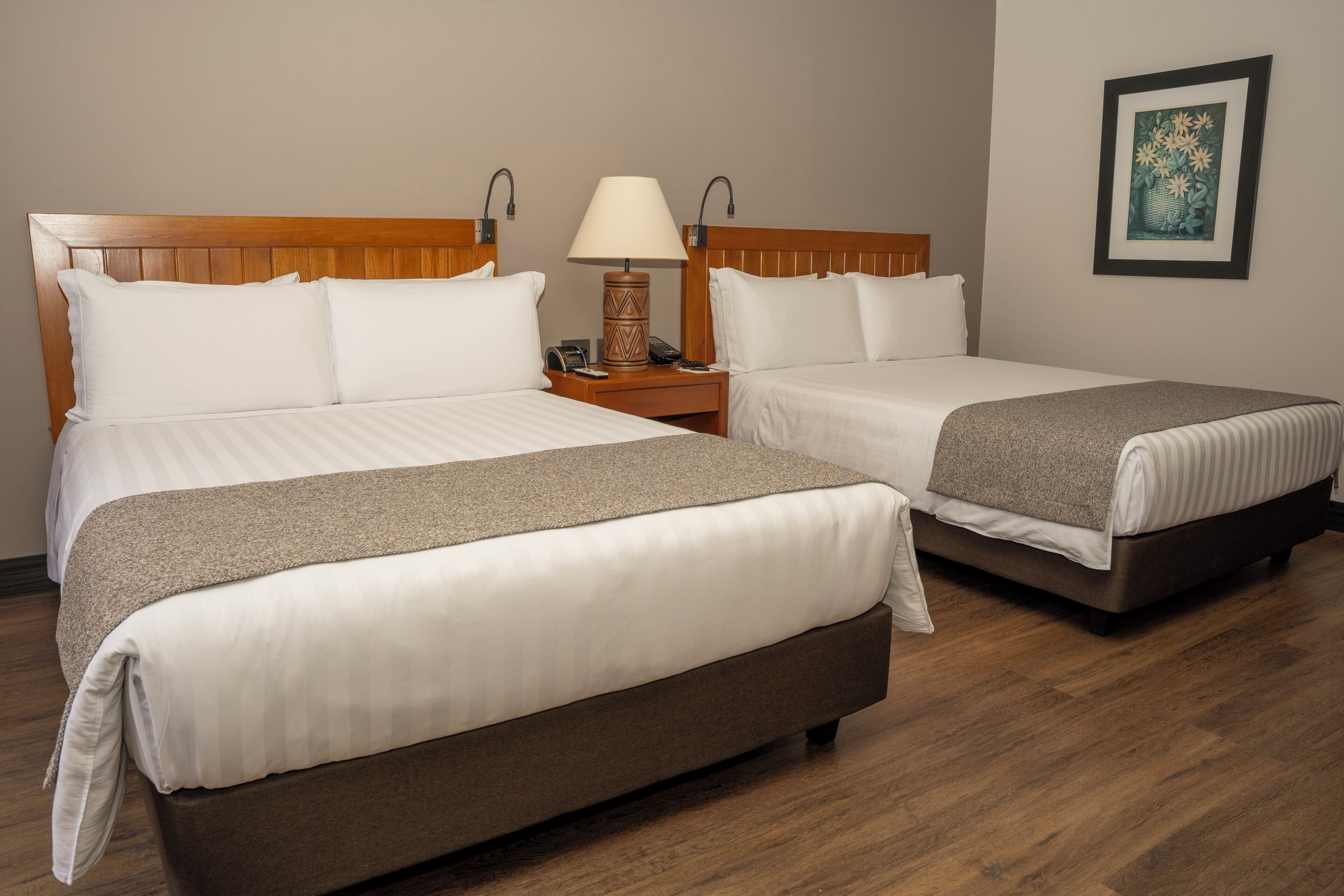 A hotel room with two beds, a nightstand with a lamp, a framed picture on the wall, and wooden flooring.