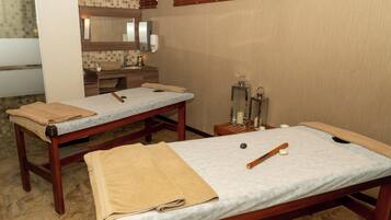 A spa room with two massage tables, towels, a wooden bench, a mirror, and a wall-mounted massage chair.