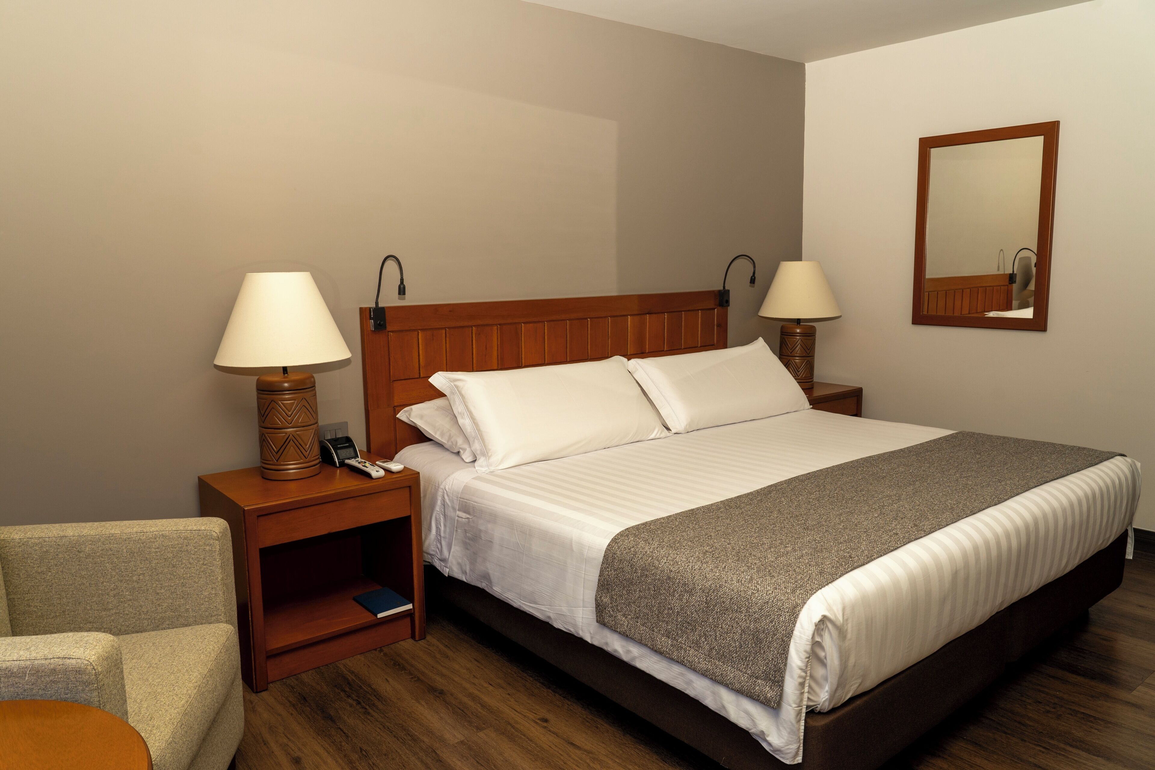 Quarto de hotel com cama, duas luminárias de cabeceira, mesa de apoio, cadeira e espelho na parede.