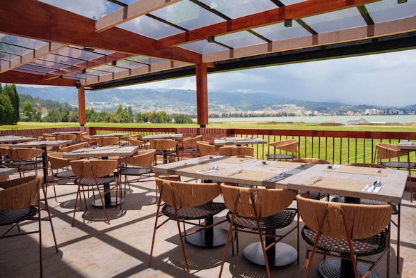 An outdoor dining area with tables and chairs, a glass ceiling, and a view of a town and mountains in the background. - Estelar Paipa Hotel Spa & Centro de Convenciones (Paipa)