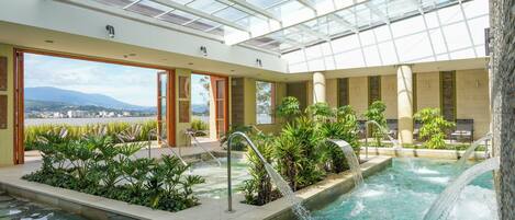 An indoor swimming pool with a glass ceiling, surrounded by plants and seating areas.