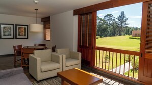A living room with a sofa, armchairs, a coffee table, a dining table with chairs, a lamp, and a large window with a view of a lawn and trees. - Estelar Paipa Hotel Spa & Centro de Convenciones (Paipa)