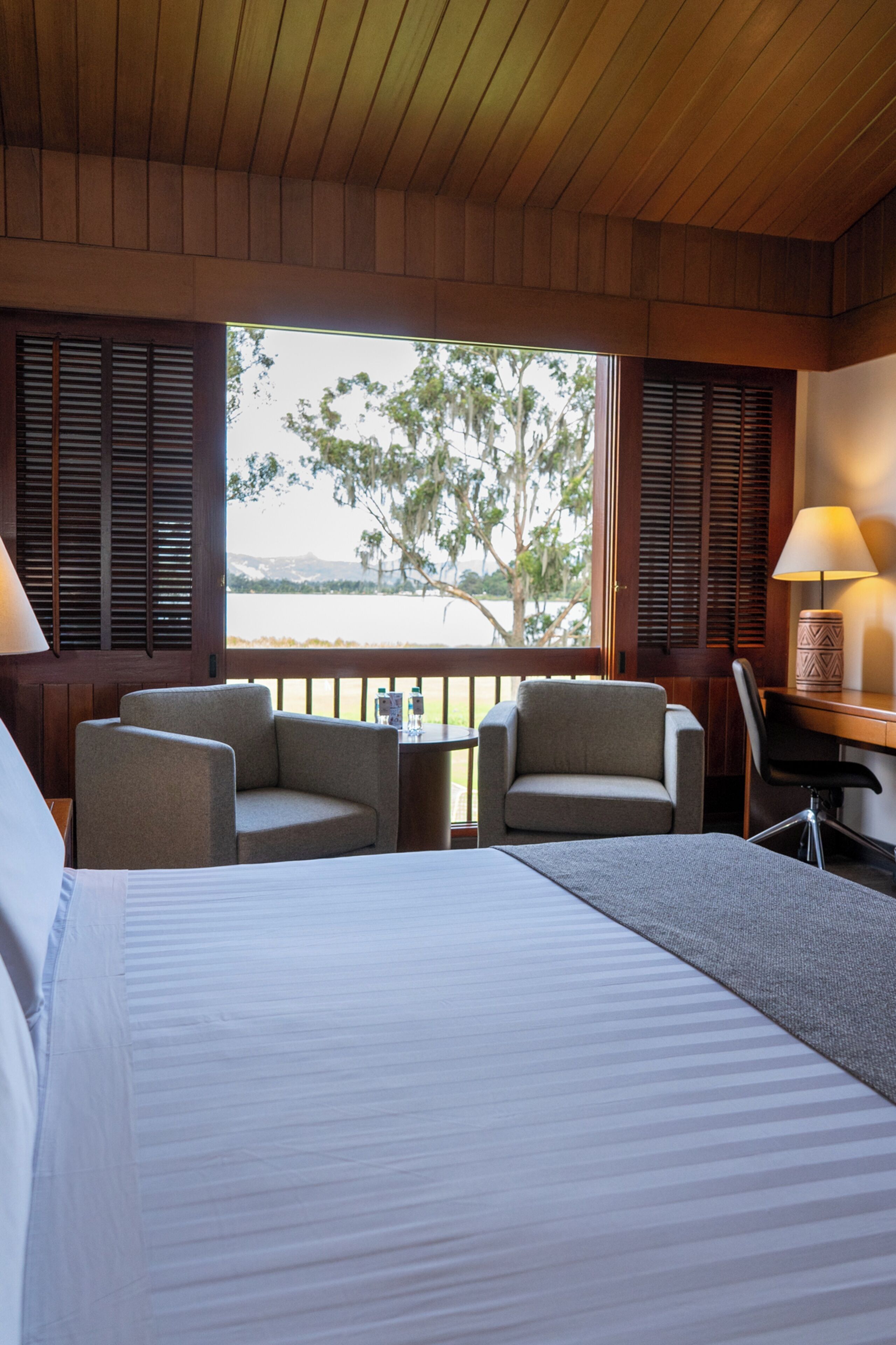 A hotel room with a bed, two armchairs, a desk, and a view of a body of water.