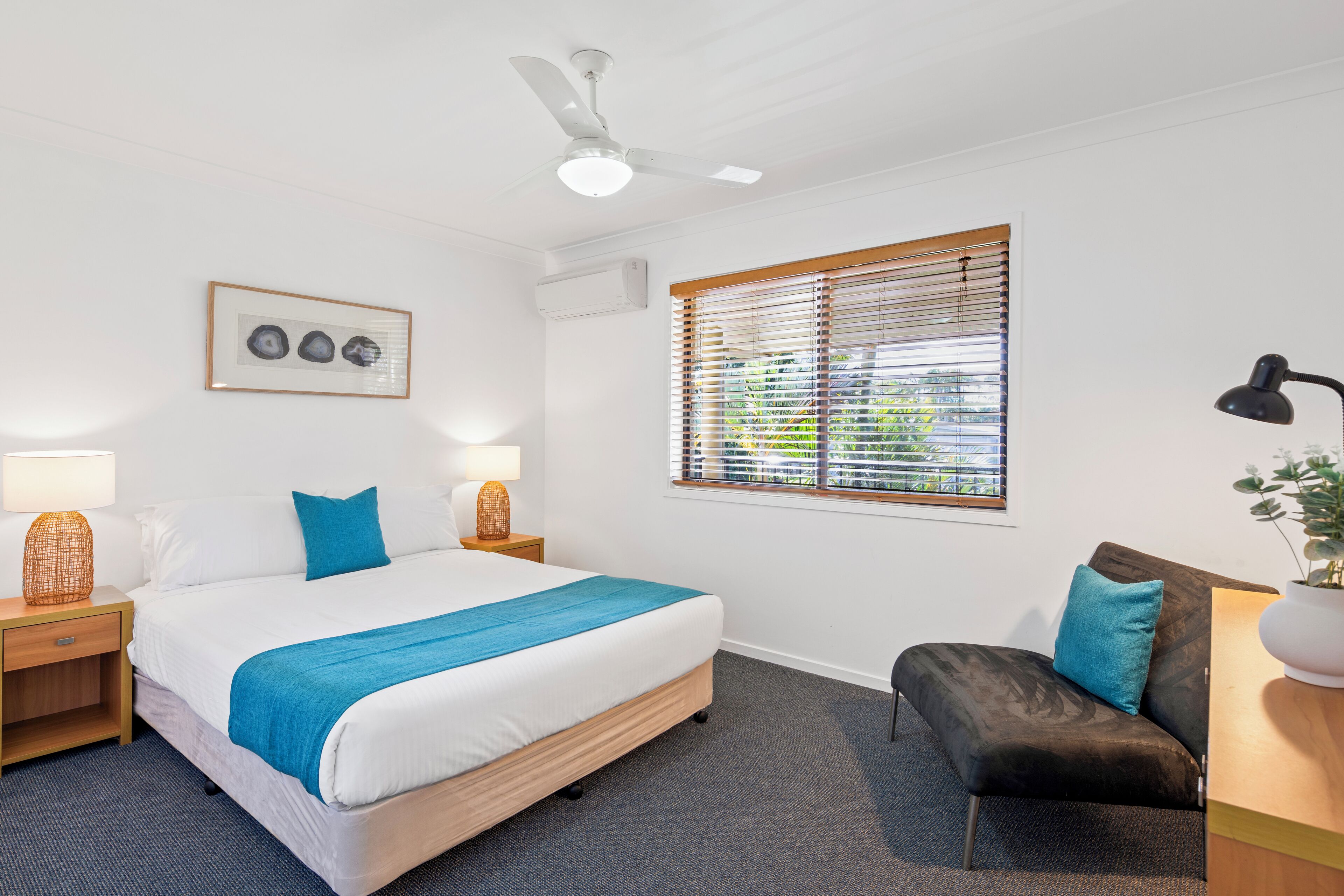 A hotel room with a bed, a desk, a chair, a ceiling fan, a window with blinds, and a framed picture on the wall.