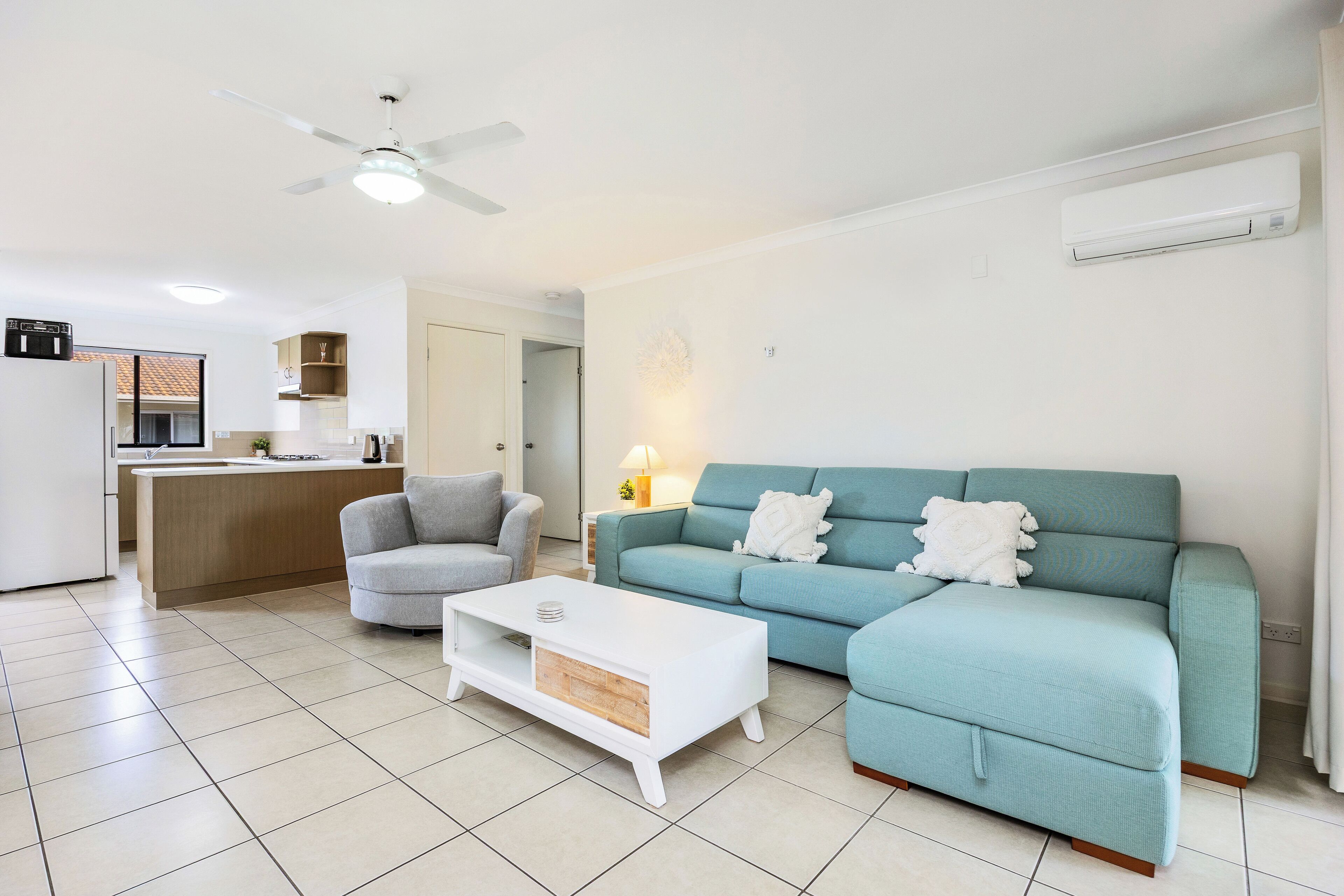 A modern living room with a sofa, armchair, and coffee table, a kitchen area with a refrigerator and cabinets, and a ceiling fan.