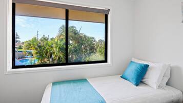 A bedroom with a bed, a window with a view of a swimming pool and palm trees, and a white wall.