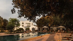 A pool area with lounge chairs and umbrellas, surrounded by trees and a building with multiple windows. - Hotel Rural Sant Ignasi (Ciutadella de Menorca)
