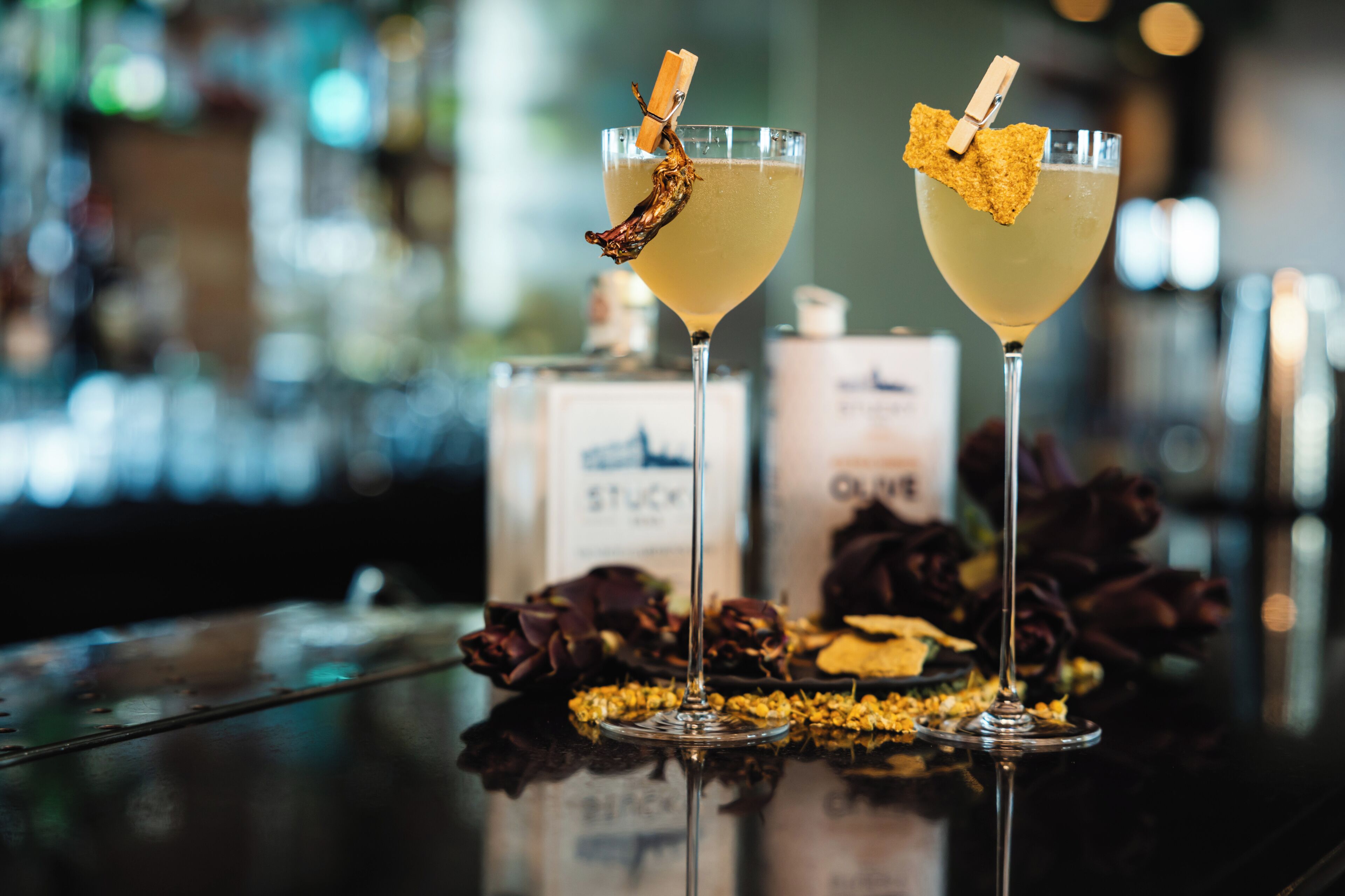 Two glasses of a yellow beverage with a dried flower garnish on a reflective surface, with bottles and flowers in the background.