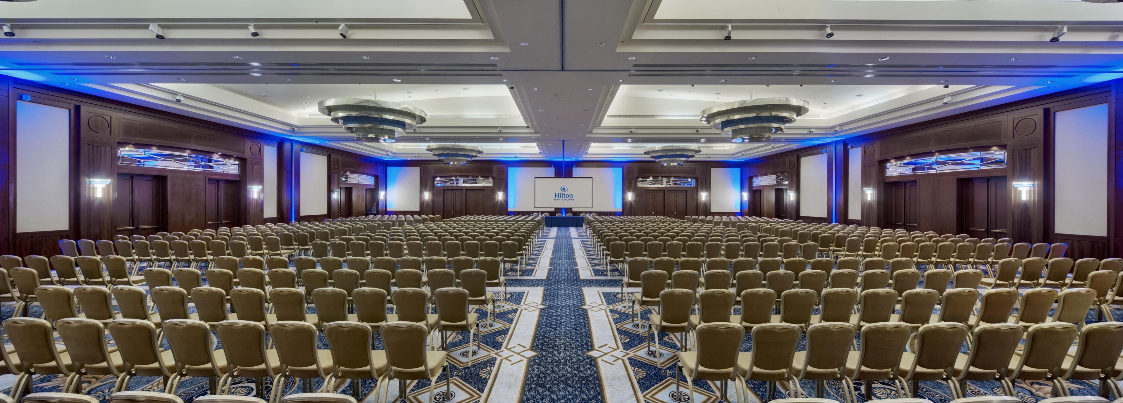 Un salón de conferencias amplio con filas de sillas, un escenario y una pantalla que muestra el logotipo de Hilton.