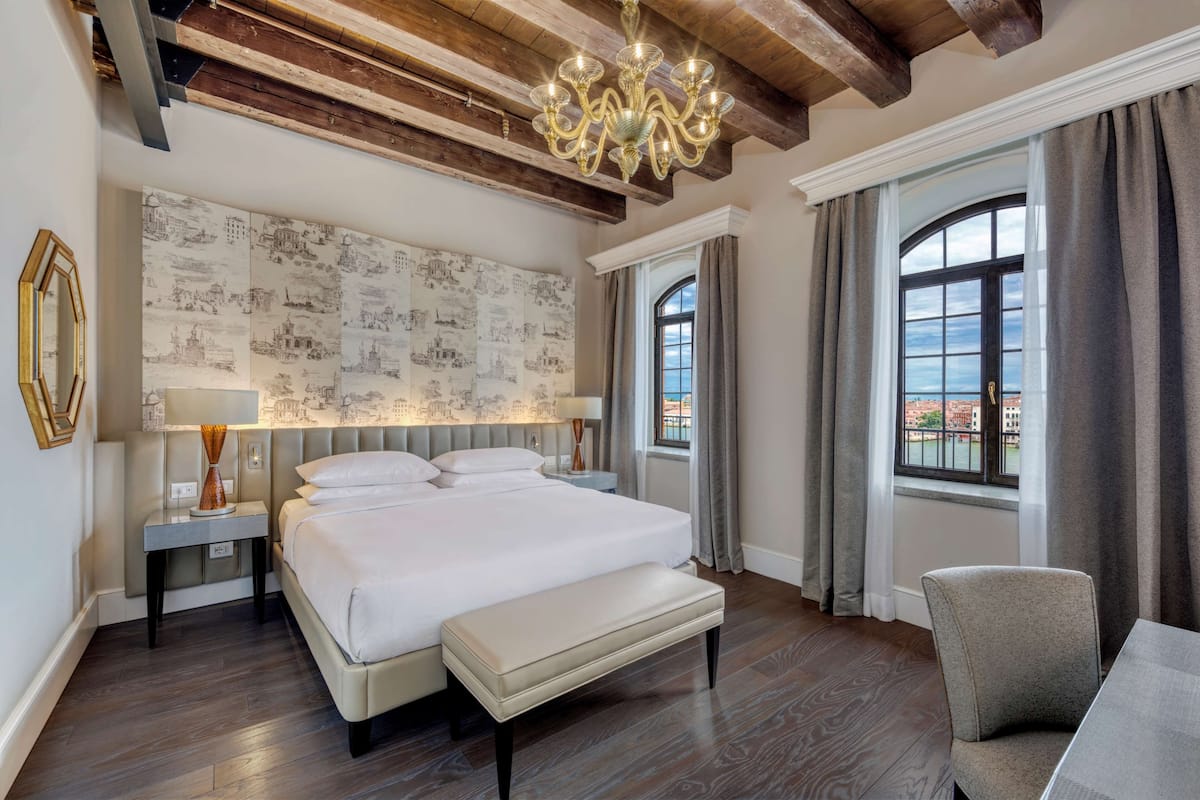 Une chambre d’hôtel moderne avec un grand lit, une banquette, une chaise et une vue sur la ville par la fenêtre.
