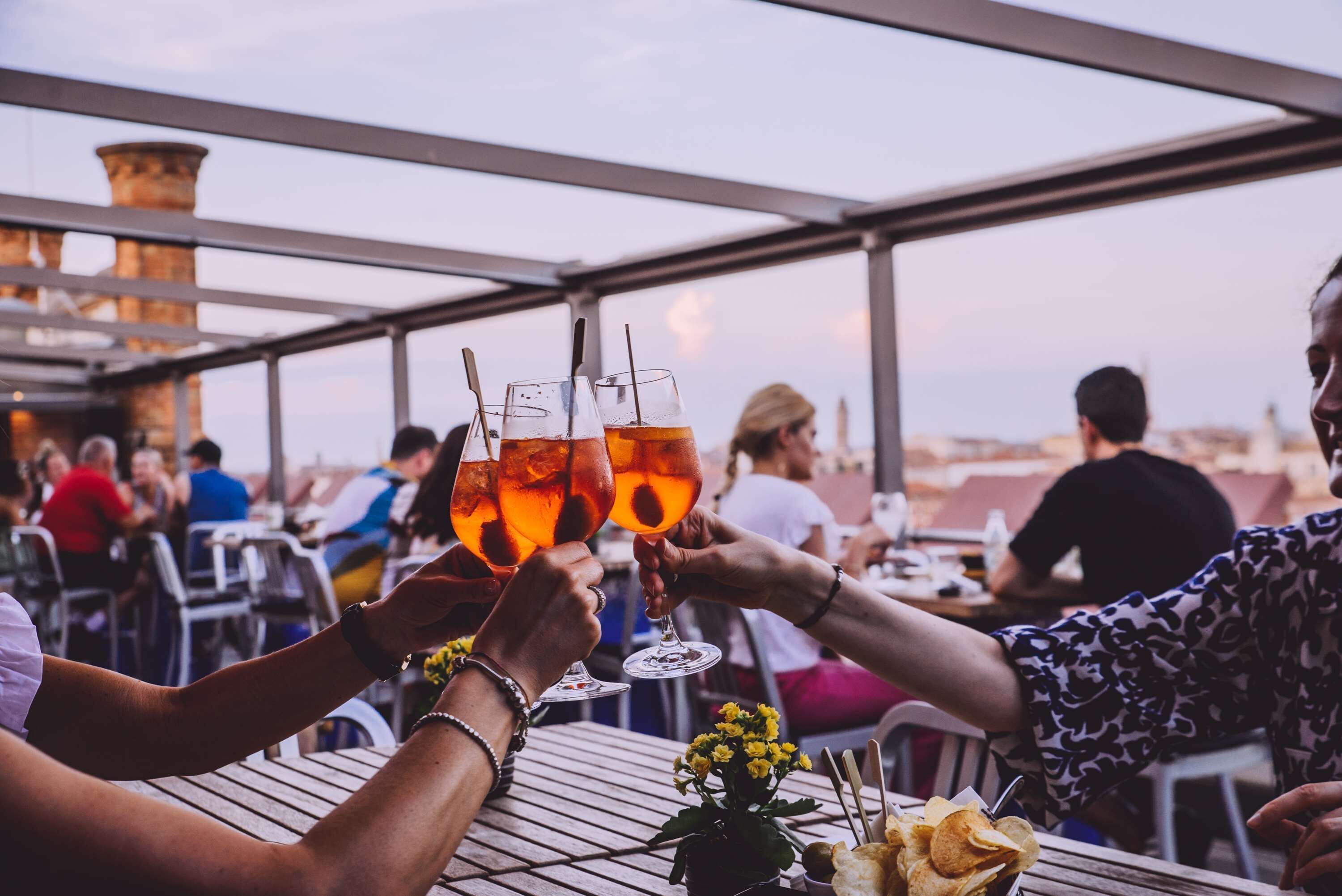 Un grupo de personas brindando con bebidas en una mesa al aire libre, con edificios al fondo.