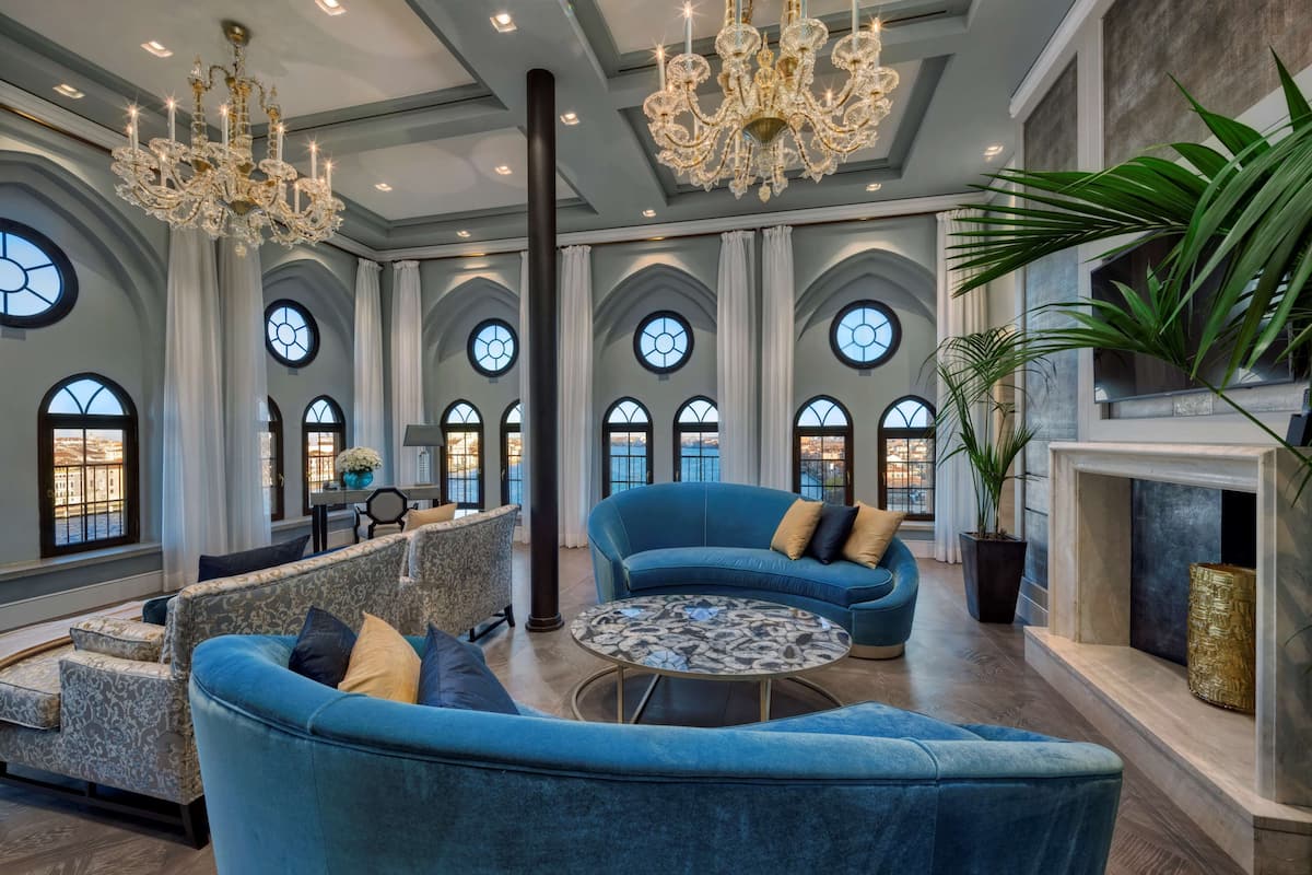 Une pièce spacieuse avec un grand canapé bleu, une table basse ronde et une cheminée. De nombreuses fenêtres à arc en plein cintre illuminent la pièce, et un lustre est suspendu au plafond.
