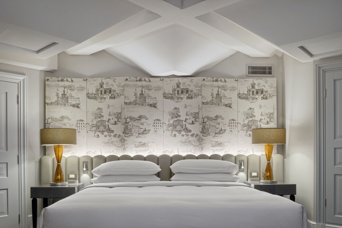 Camera d'albergo con un letto grande, due comodini con lampade e una parete con carta da parati raffigurante un paesaggio urbano.
