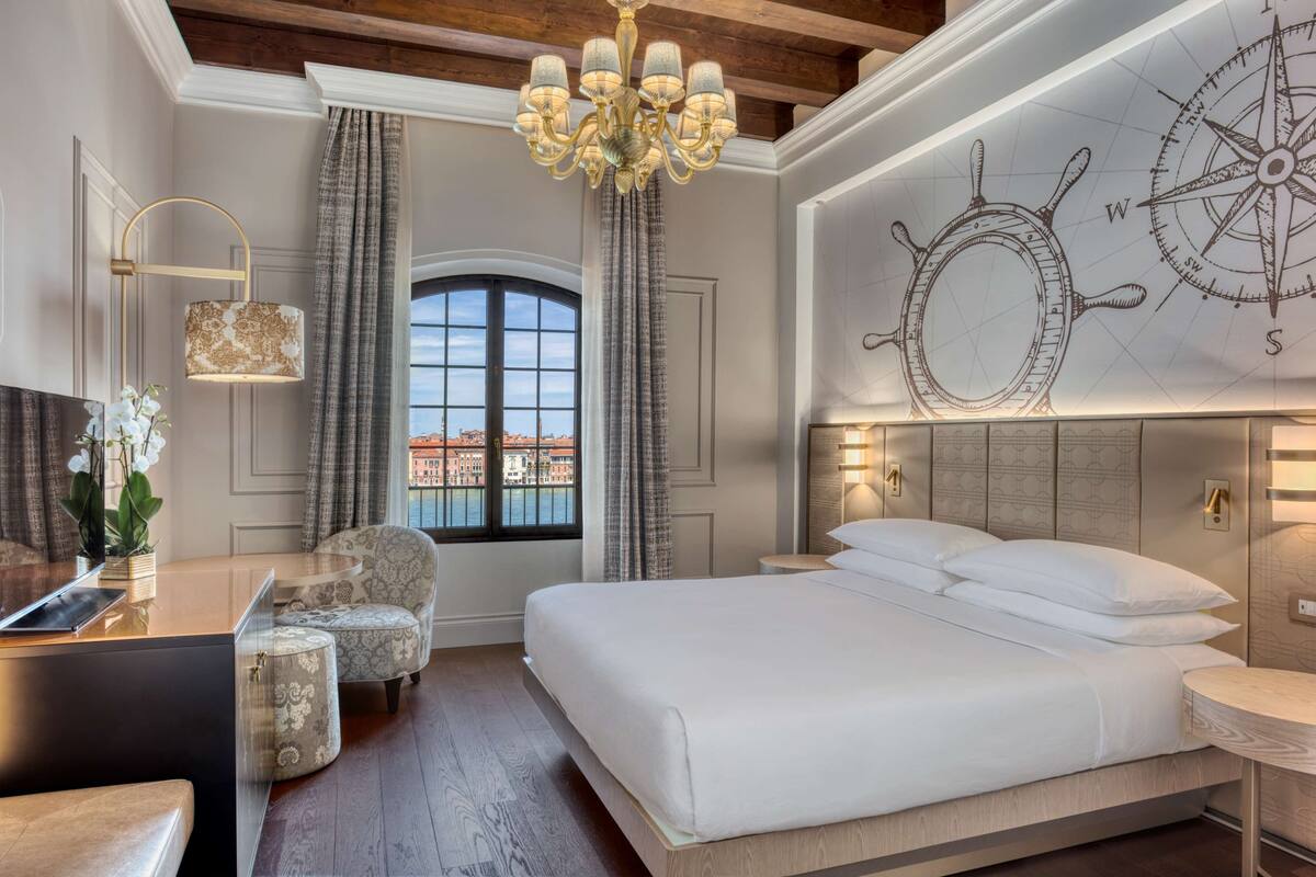 Une chambre d’hôtel avec un grand lit, un bureau avec une lampe, une chaise, une vue sur le paysage urbain par la fenêtre et une carte murale.