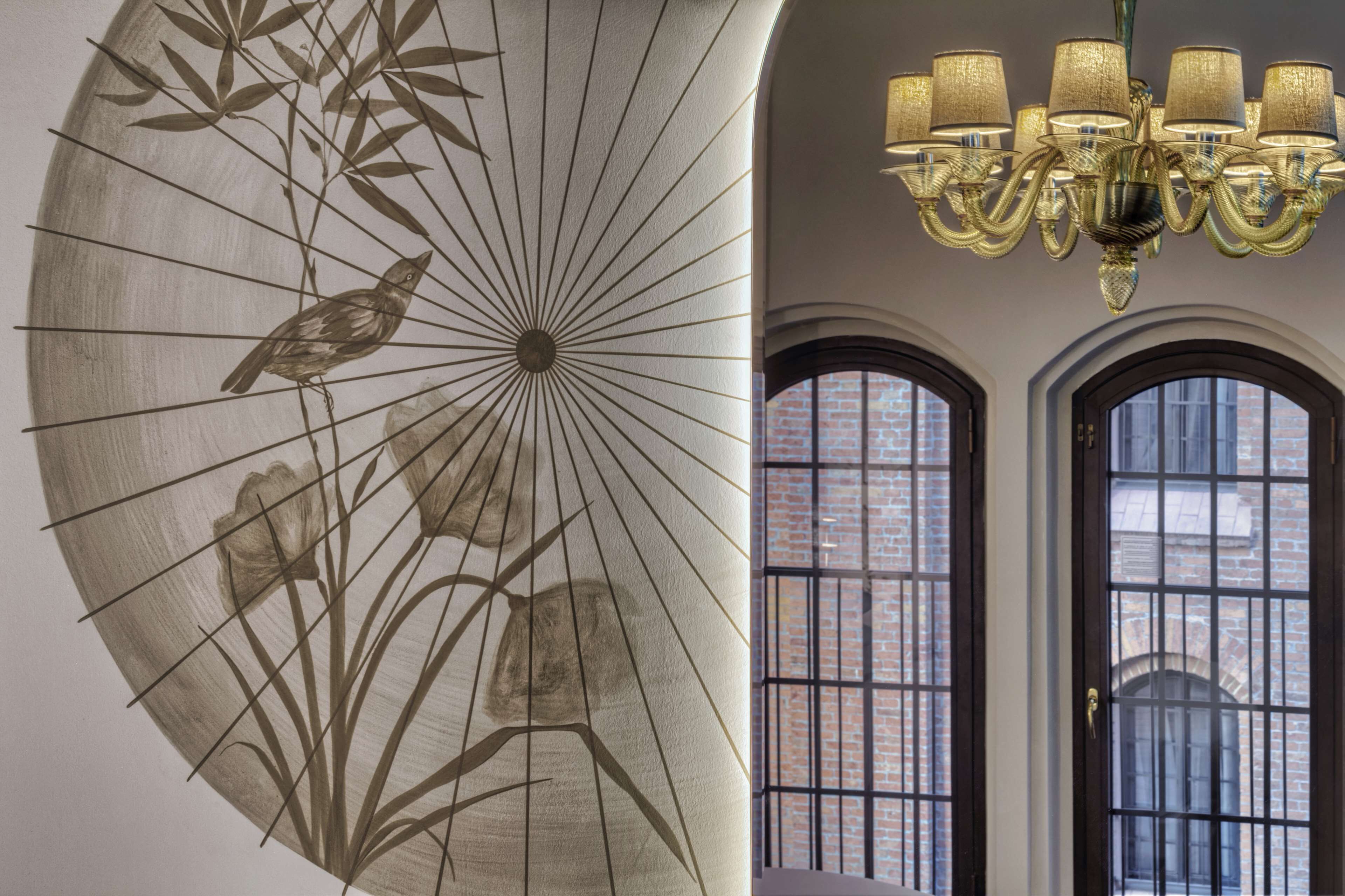 A room with a large wall mural of bamboo and a bird, a chandelier, and two arched windows with a brick wall visible through them.