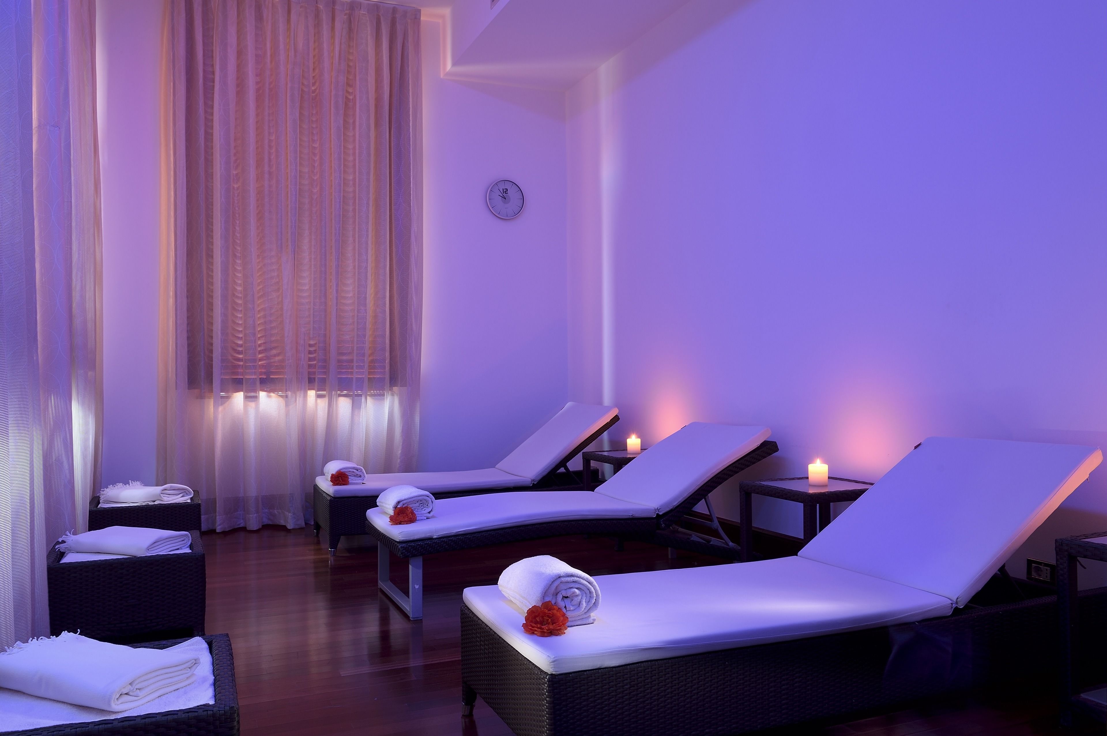 Une salle de spa équipée de deux tables de massage, chacune agrémentée d’une serviette et d’une fleur. Une fenêtre est ornée de rideaux légers et une horloge est accrochée au mur.