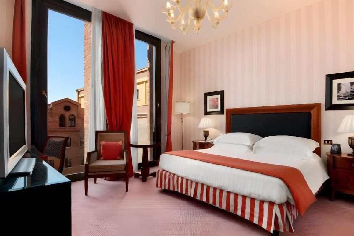 A hotel room with a large bed, a desk, a chair, a TV, and a view of buildings outside.