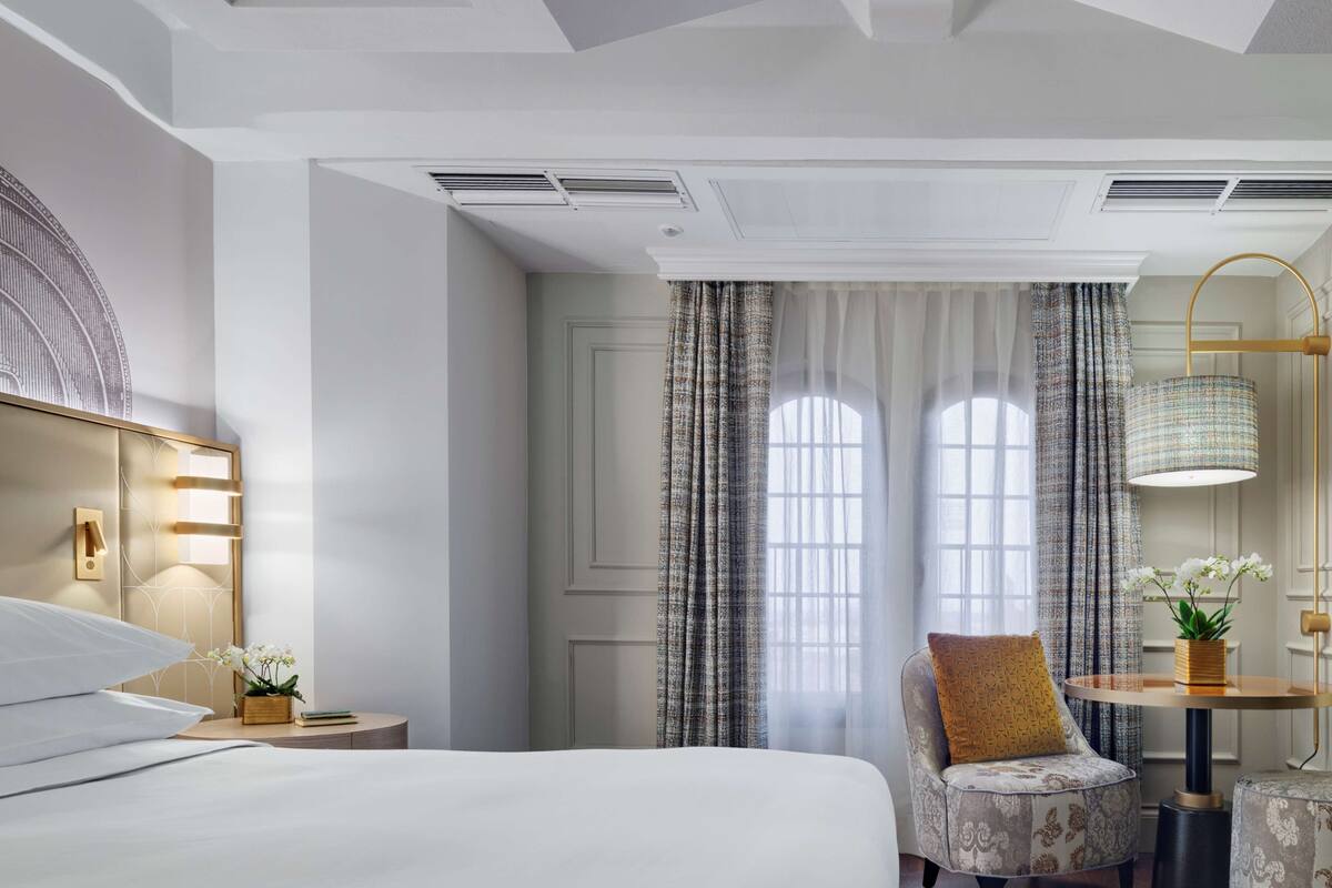 Une chambre d’hôtel avec un lit, une chaise, une table avec une lampe et une fenêtre avec des rideaux.