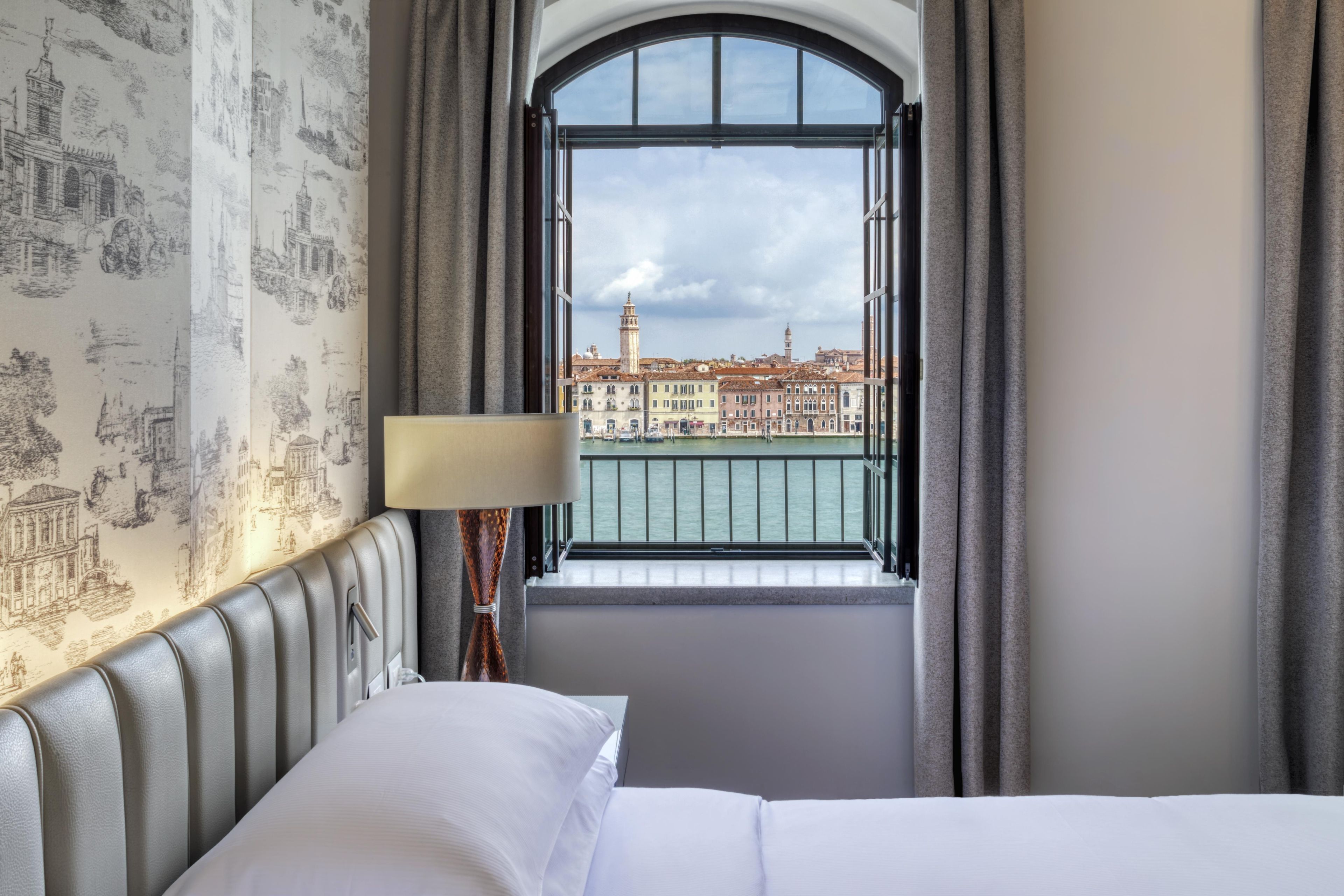 Une chambre d’hôtel avec un lit, une fenêtre avec des rideaux, une lampe et une vue sur le paysage urbain.