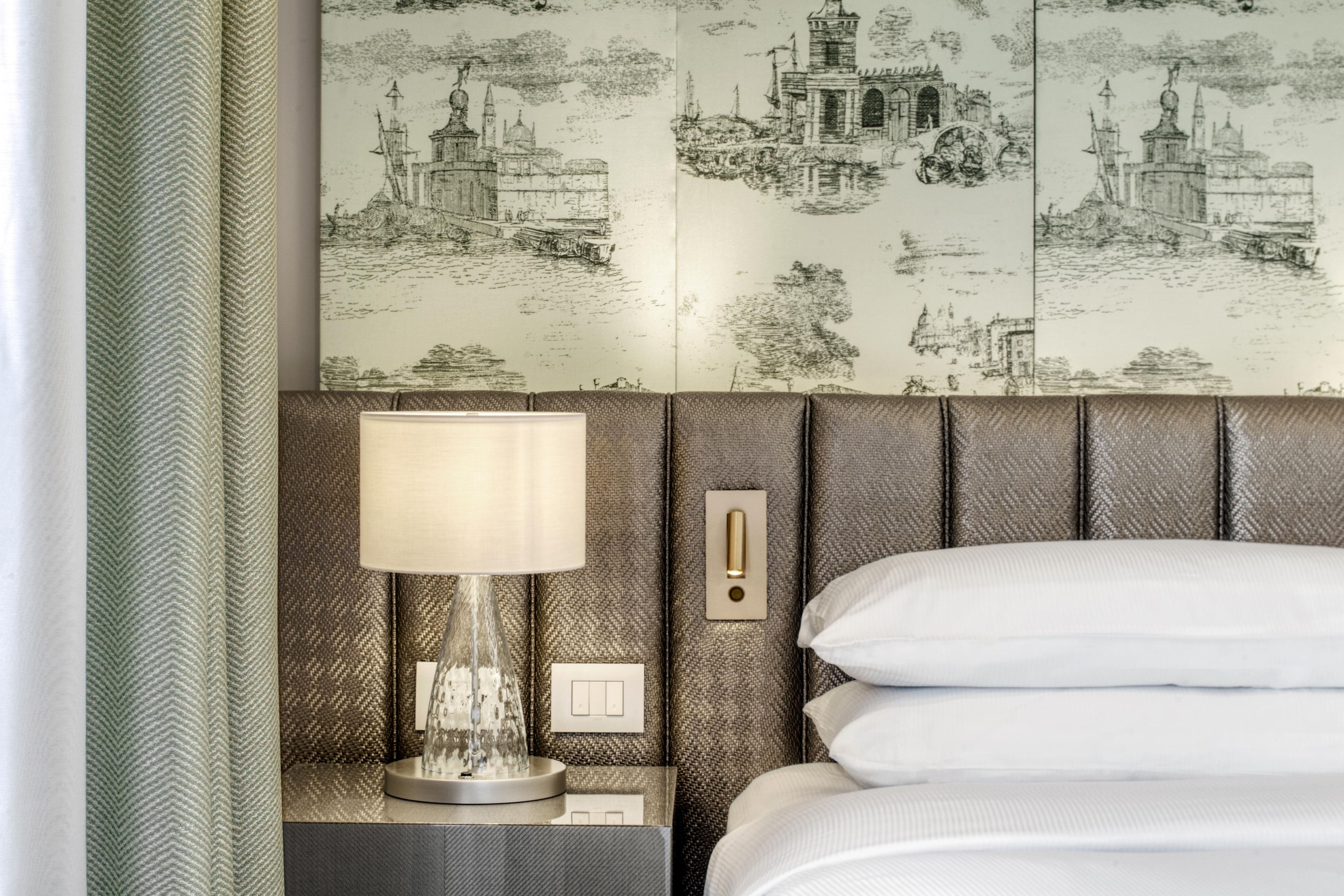 Habitación de hotel con una cama, una mesita de noche con lámpara y una pared con un mural de paisaje urbano.