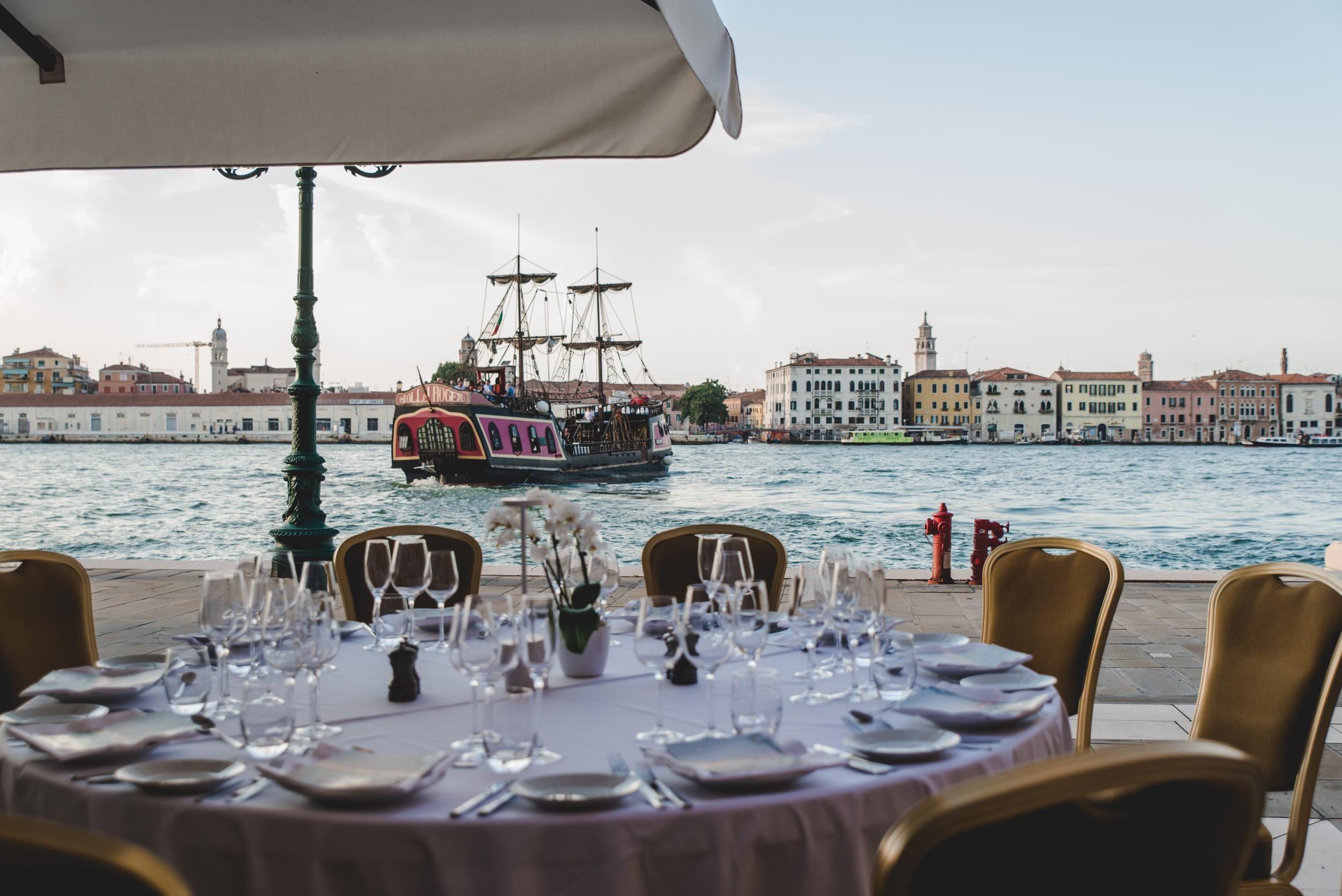 A waterfront dining table with chairs, a ship in the background, and buildings along the water.